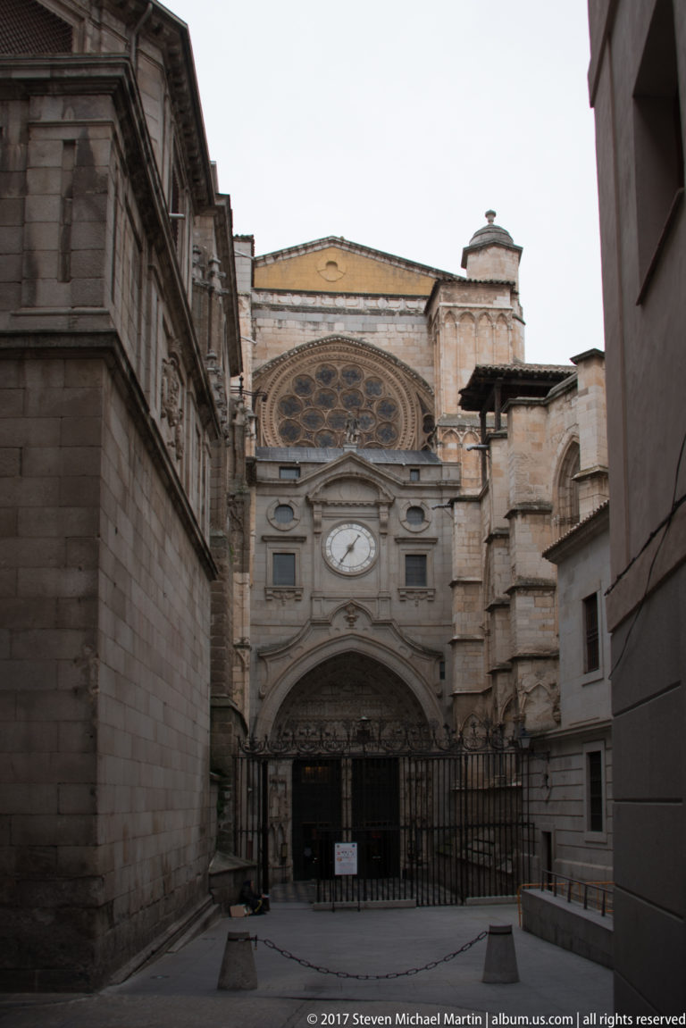 Stone church façade with arched entrance, circular window, and iron gate at the end of a narrow Toledo urban space.