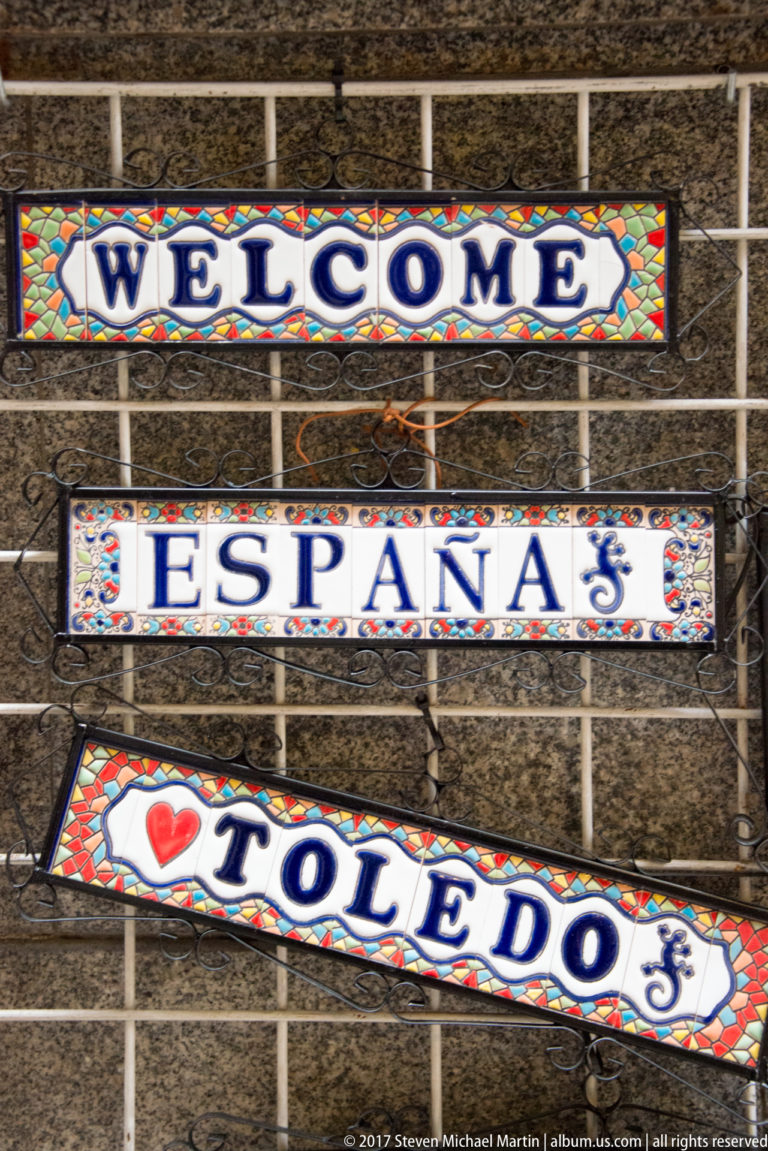 Three colorful decorative signs reading “WELCOME,” “ESPAÑA,” and “I love TOLEDO” mounted on a metal grid.