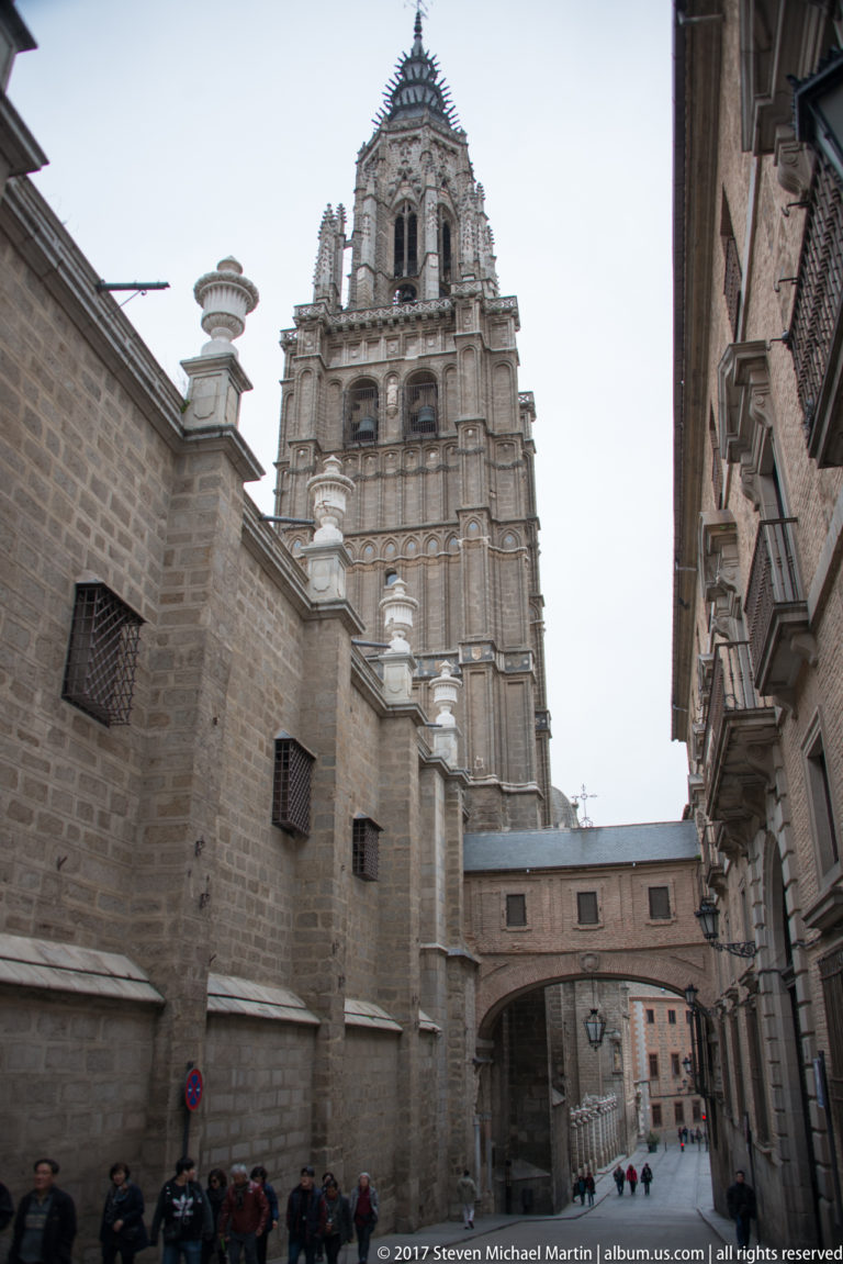 Gothic bell tower of Toledo Cathedral rising above a narrow stone-lined street with pedestrians and an overhead passage.