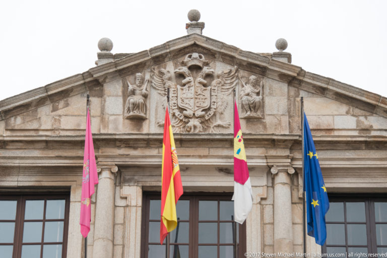 Stone civic façade with carved heraldic eagle and shield above Spanish, regional, local, and European Union flags.