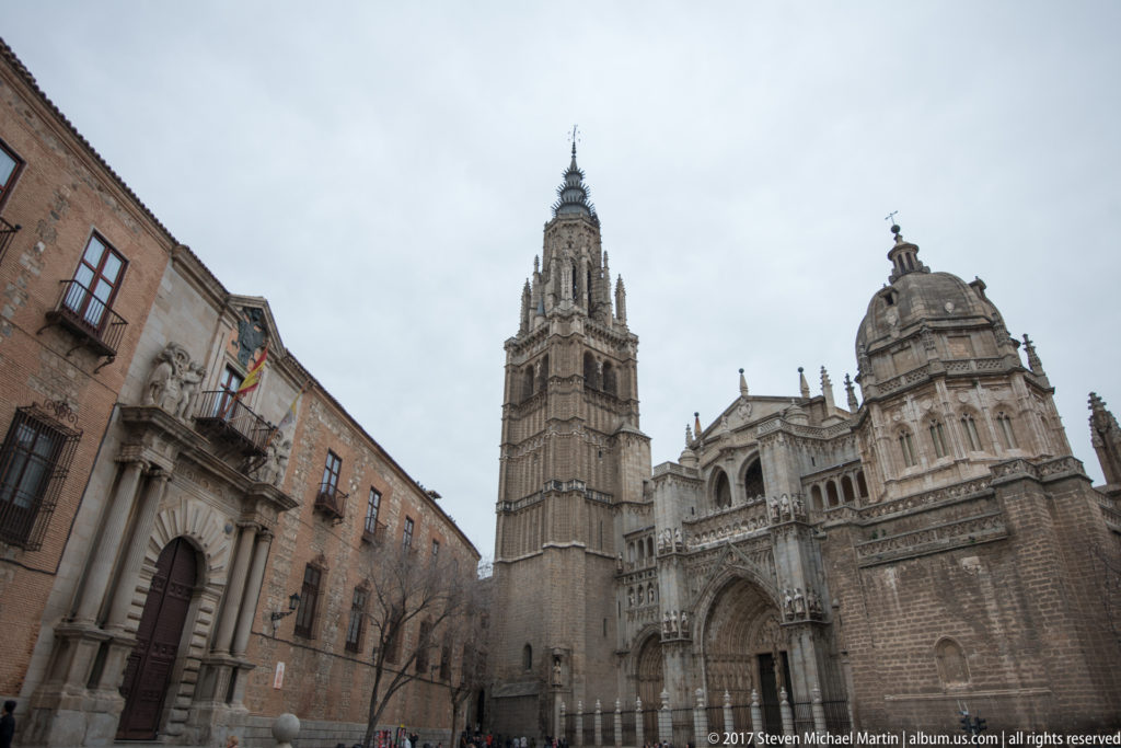 Toledo Cathedral seen from below with its tall Gothic bell tower, sculpted entrance façade, and adjoining domed structure.