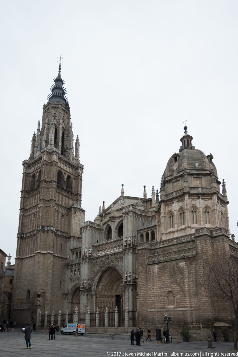 Wide exterior view of Toledo Cathedral showing the Gothic bell tower, sculpted entrance, and rounded adjoining structure.