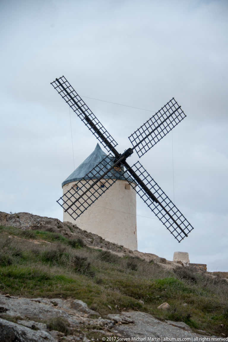 White cylindrical tower windmill with lattice sails and a dark conical roof on a rocky ridge under cloudy sky.