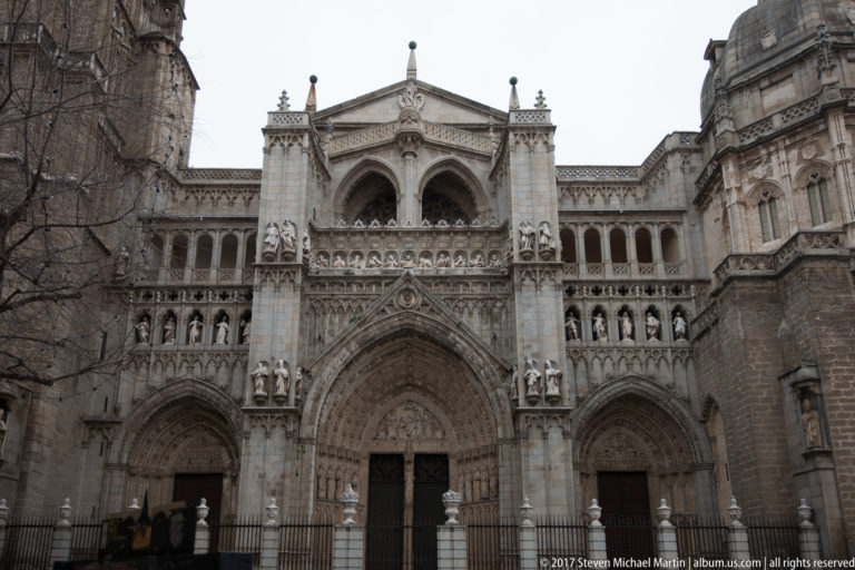 Frontal view of an ornate Gothic cathedral portal with pointed arches, rows of sculptures, tracery, and iron fencing.