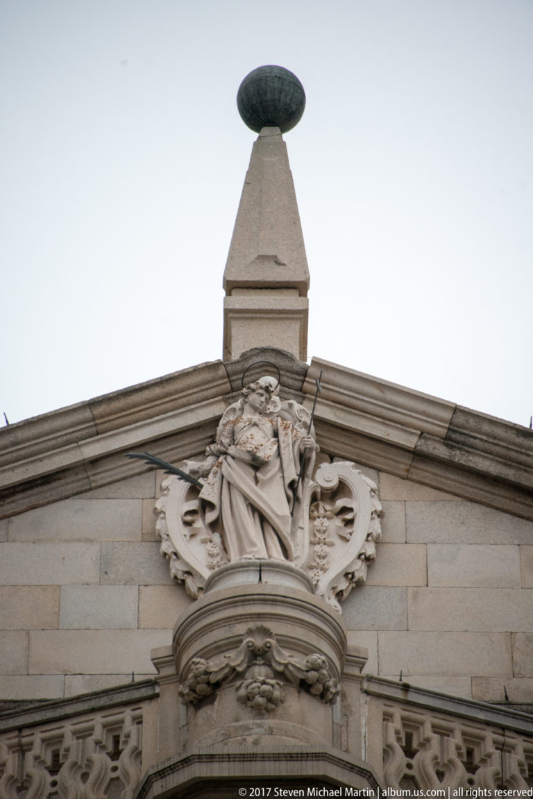 Carved robed figure set high in an ornate stone pediment beneath a finial and oval ornament.