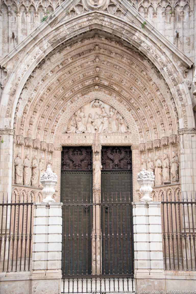 Gothic cathedral doorway with multiple carved archivolts, standing figures, narrative tympanum, and paired dark doors.