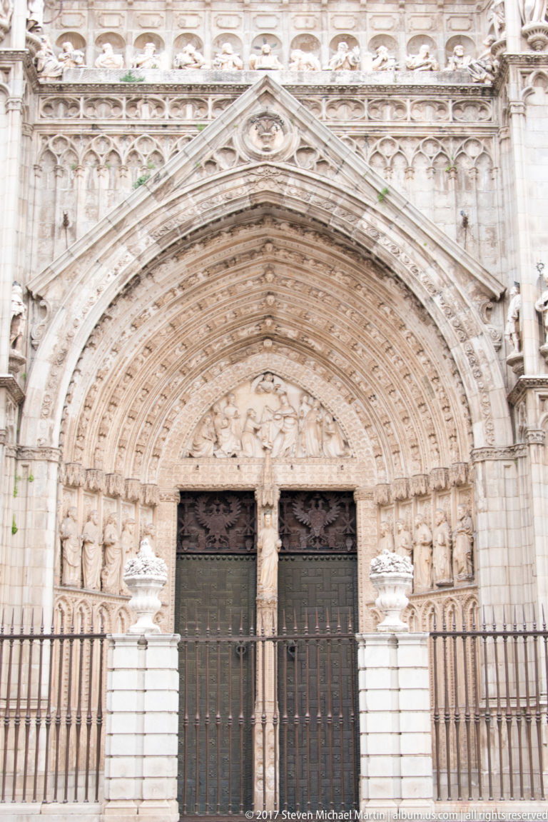 Frontal view of a Toledo Cathedral portal with carved archivolts, sculpted figures, paired doors, and iron railings.