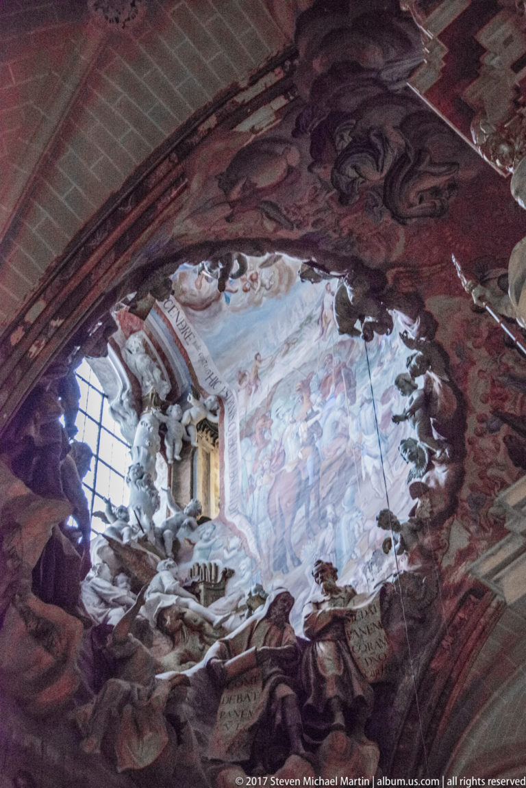Oblique view of an illusionistic ceiling fresco with clouds, figures, painted architecture, and dark red-brown framing.