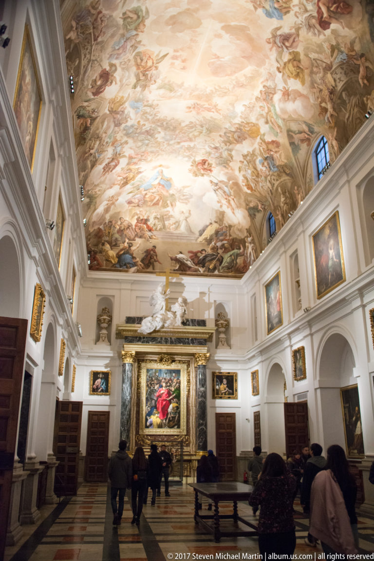 Long cathedral room with a large painted ceiling, framed artworks, dark wooden furnishings, and visitors gathered near a monumental end display.