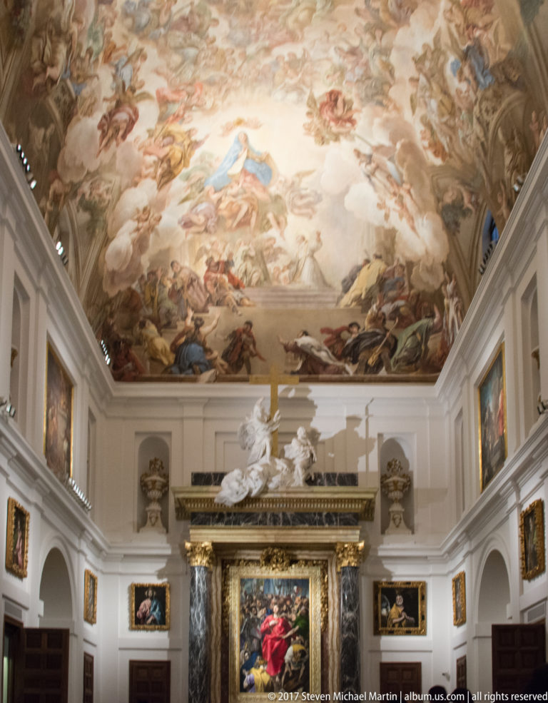 Upward view of a cathedral room with a luminous frescoed ceiling, sculptural group, and large framed painting on the end wall.
