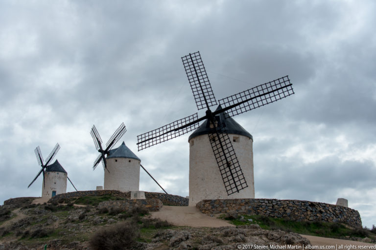 Three white tower windmills with lattice sails spaced along a rocky ridge beneath dark clouds.