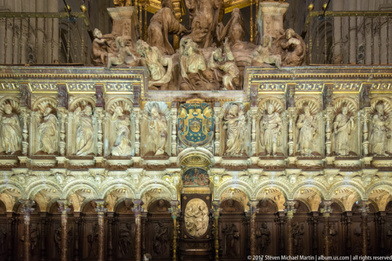Ornate cathedral screen with rows of carved figures, gilded reliefs, a central heraldic shield, and dark woodwork below.