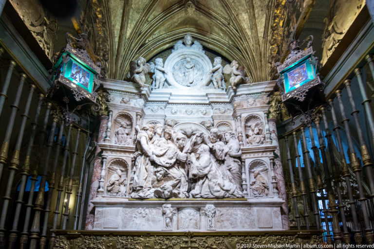 Large pale-stone cathedral monument with a central multi-figure relief, smaller panels, sculpted figures, and a circular emblem above.
