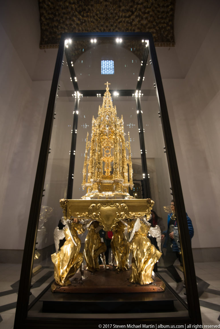 Tall gilded liturgical object in a glass case, formed as intricate miniature architecture above a sculptural base.