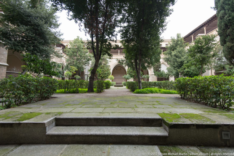 Landscaped cloister courtyard with stone paving, clipped hedges, mature trees, and arched galleries around the garden.
