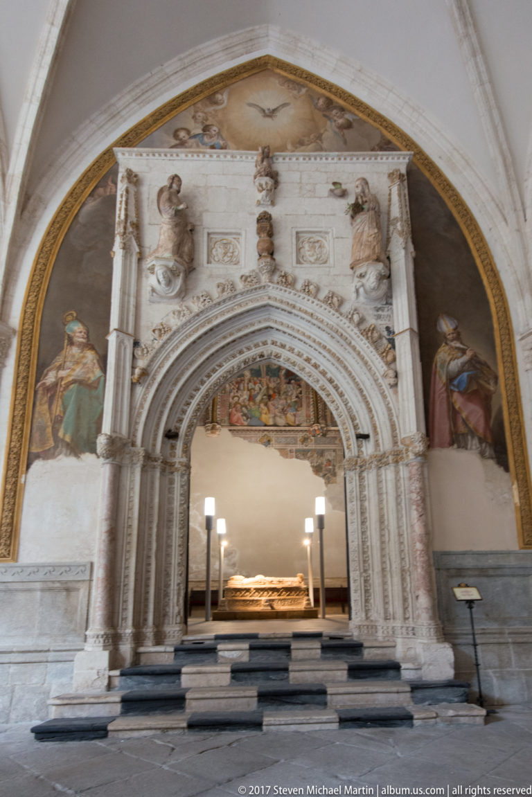 Ornate stone portal with painted saints and carved figures opening toward an illuminated recumbent tomb on a raised platform.