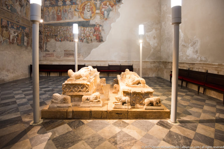 Two recumbent stone effigies on adjacent tomb chests, illuminated against a wall with surviving historic painting.