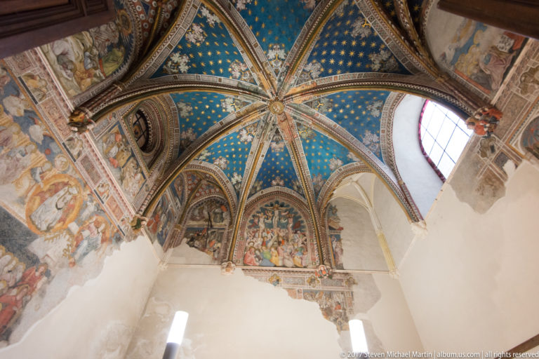 Blue and gold ribbed vault above fragmentary figurative wall paintings in a small historic cathedral interior.