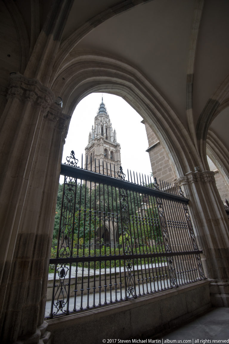 Toledo Cathedral tower seen through a broad stone cloister arch and iron grille above green foliage.