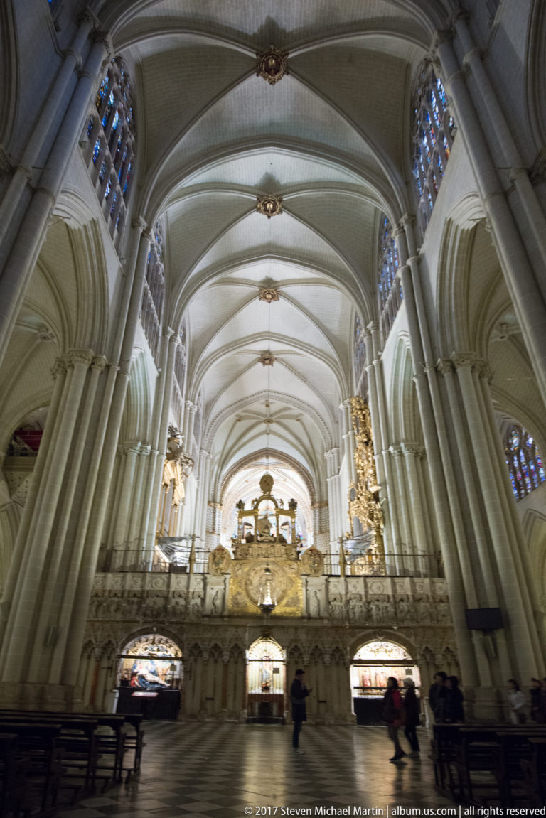 Interior of Toledo Cathedral with tall clustered columns, ribbed vaults, stained glass, and an ornate enclosure across the nave.