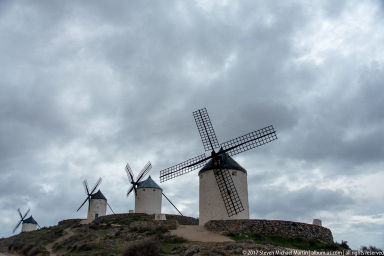 Several white tower windmills recede along a ridge under heavy gray clouds, with the largest mill at right.