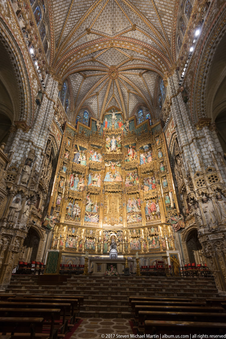Wide interior view of Toledo Cathedral’s gilded high retablo beneath ribbed vaulting, framed by massive piers and seating.