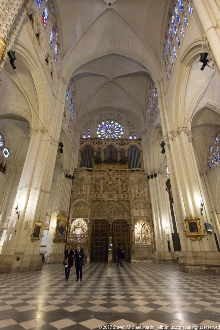 Wide Toledo Cathedral interior with pale Gothic vaults, organ case, rose window, patterned floor, and a few visitors.
