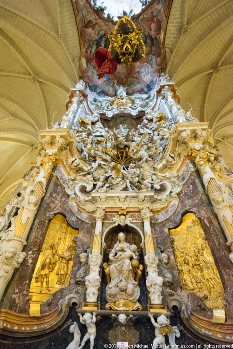 Upward frontal view of the Transparente combining marble altar architecture, pale sculpture, painted clouds, and a bright opening overhead.