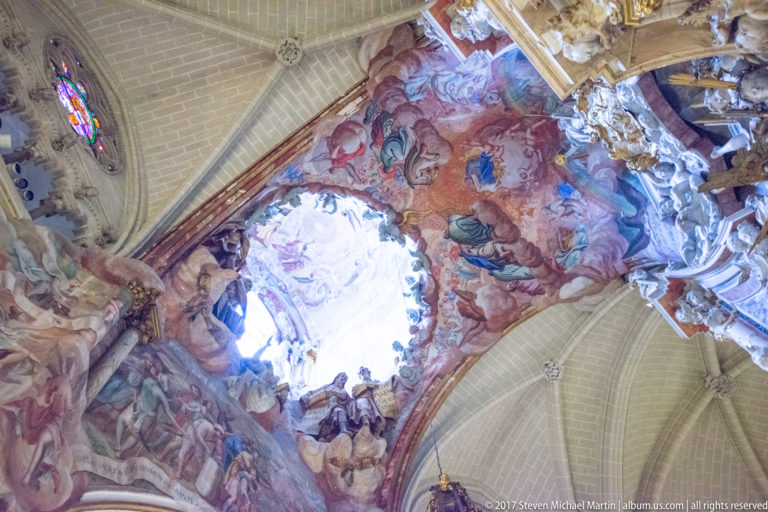 Upward view of the Transparente’s bright opening surrounded by painted figures, clouds, sculpture, and adjacent Gothic vaulting.