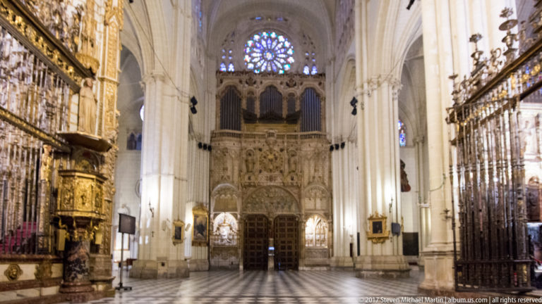 Gothic interior of Toledo Cathedral looking toward an ornate organ loft and rose window, with screens and patterned stone floor.