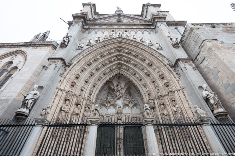 Low-angle view of a Gothic cathedral portal with carved archivolts, tracery, standing figures, and dark paired doors.