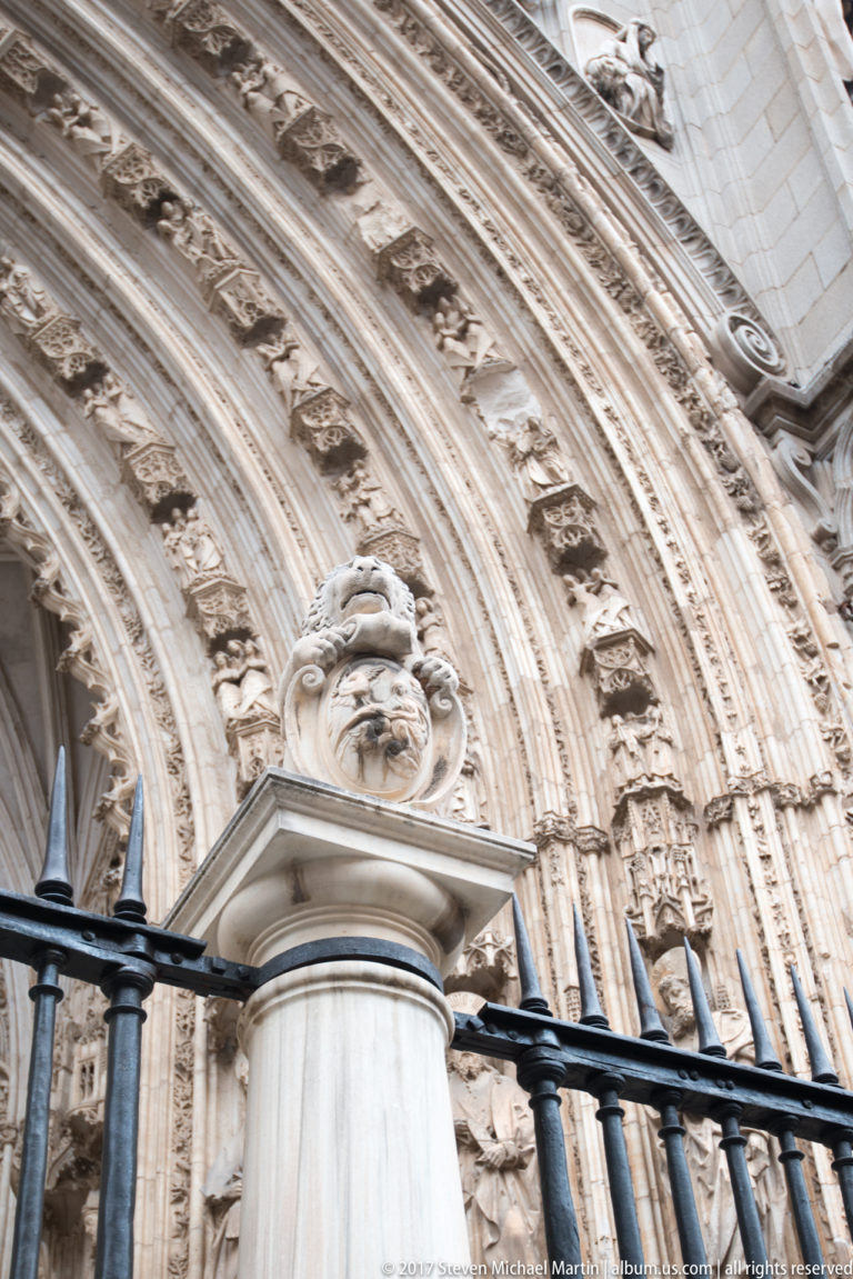 Oblique close view of carved Gothic archivolts filled with small figures above an iron fence and pale stone post.