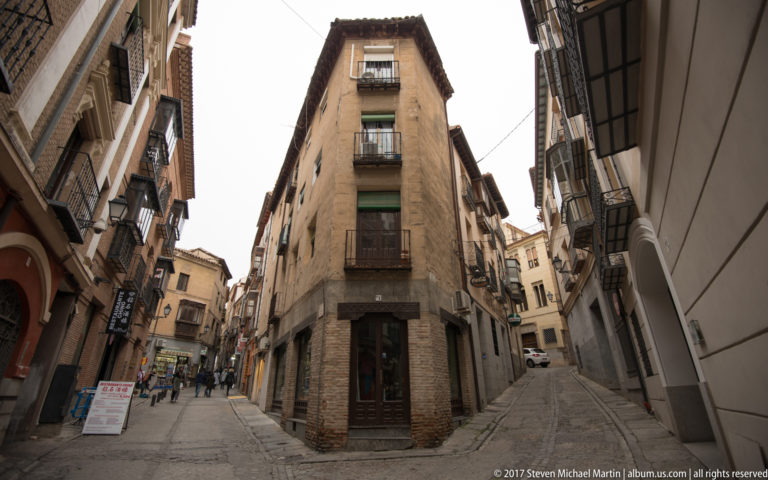 Narrow Toledo streets divide around a wedge-shaped building with balconies, brickwork, and pedestrians in the distance.