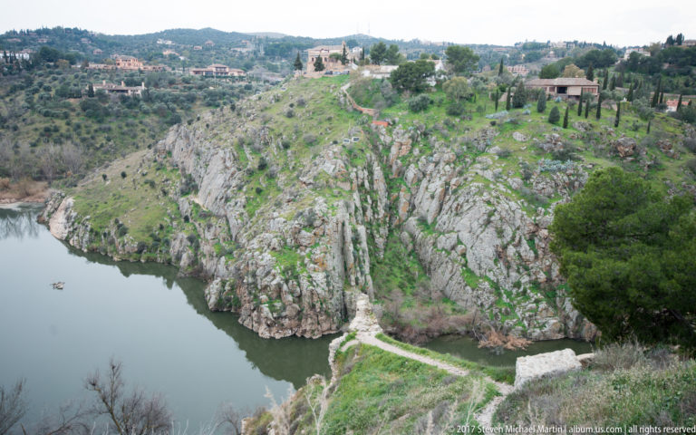 Tagus River curving through a steep rocky gorge below Toledo, with scrub vegetation, paths, and buildings on higher ground.