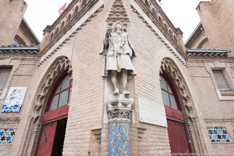 Brick corner façade with pointed arches, decorative ceramic panels, red doors, and a standing stone figure above the pier.