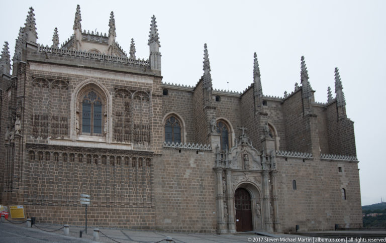 Long stone façade of San Juan de los Reyes with Gothic windows, pinnacles, tracery, and an ornate entrance portal.