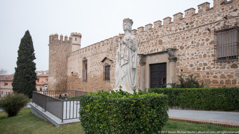 Crowned female statue beside crenellated stone walls, a square tower, hedges, and a cypress tree at San Juan de los Reyes.