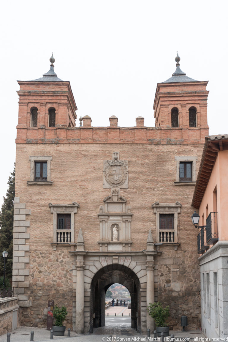 Puerta del Cambrón with central arched roadway, square corner towers, brick walls, stone trim, and a heraldic panel above.
