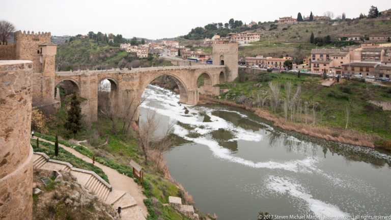 Wide view of Puente de San Martín spanning the Tagus River with its fortified tower and hillside neighborhood beyond.