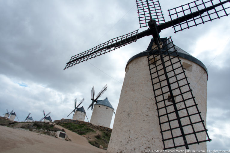 Large white windmill seen from below with diagonal lattice sails and a row of smaller mills along the ridge behind.