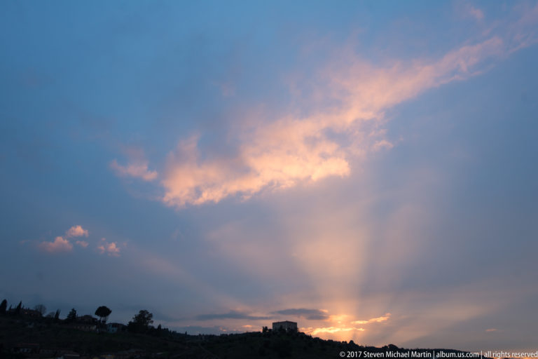 Peach-lit clouds and fan-shaped sun rays rise from the horizon above a silhouetted Toledo ridge.