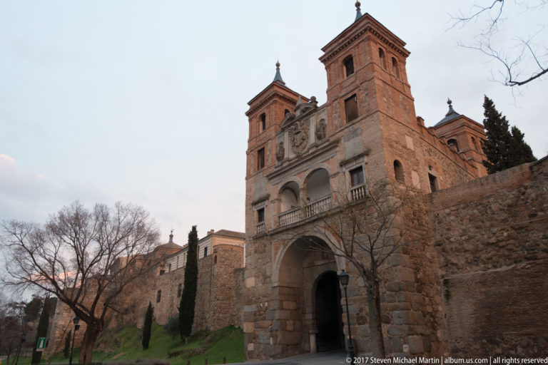 Oblique dusk view of Puerta del Cambrón with square towers, central arch, adjoining city wall, trees, and grass.