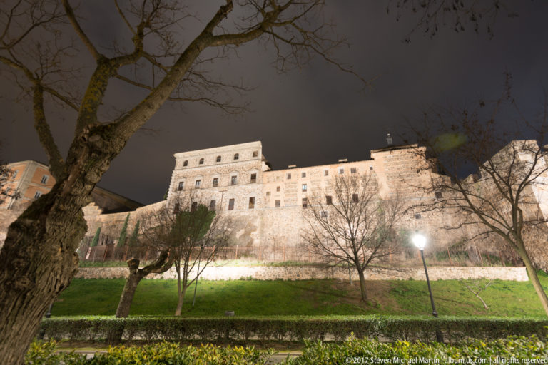 Illuminated Alcázar of Toledo above a landscaped slope with bare trees and hedges beneath a dark night sky.