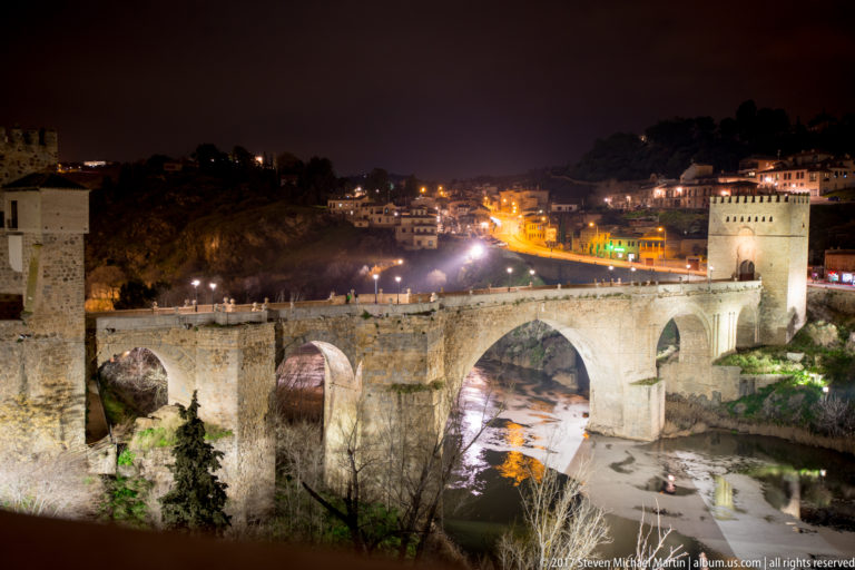 Puente de San Martín illuminated at night above the Tagus River, with glowing arches, fortified tower, and hillside lights.