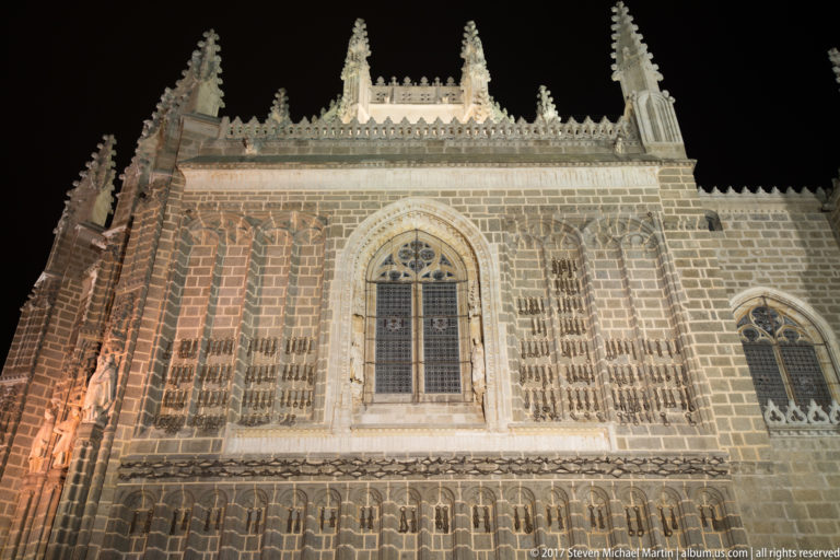 Night-lit Gothic wall of San Juan de los Reyes with traceried windows, buttresses, pinnacles, and carved roofline ornament.