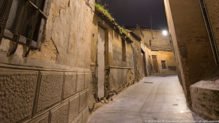Narrow Toledo alley at night with worn plaster walls, stone paving, timber windows, patterned masonry, and warm lamps.