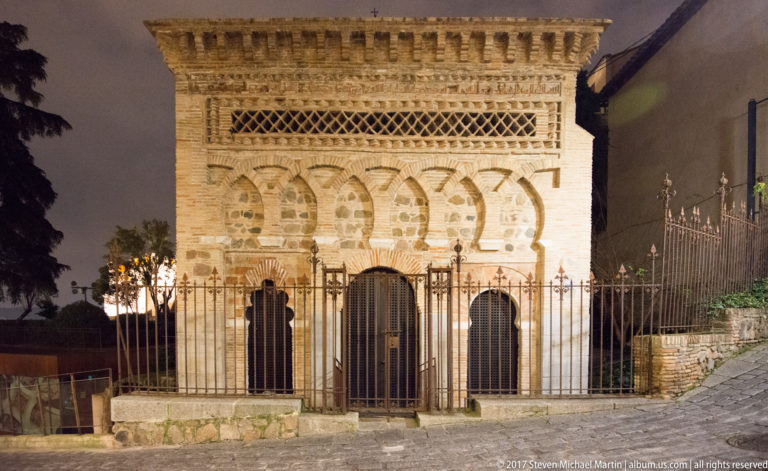 Night-lit façade of Cristo de la Luz with interlaced horseshoe arches, decorative brickwork, three lower openings, and iron fence.