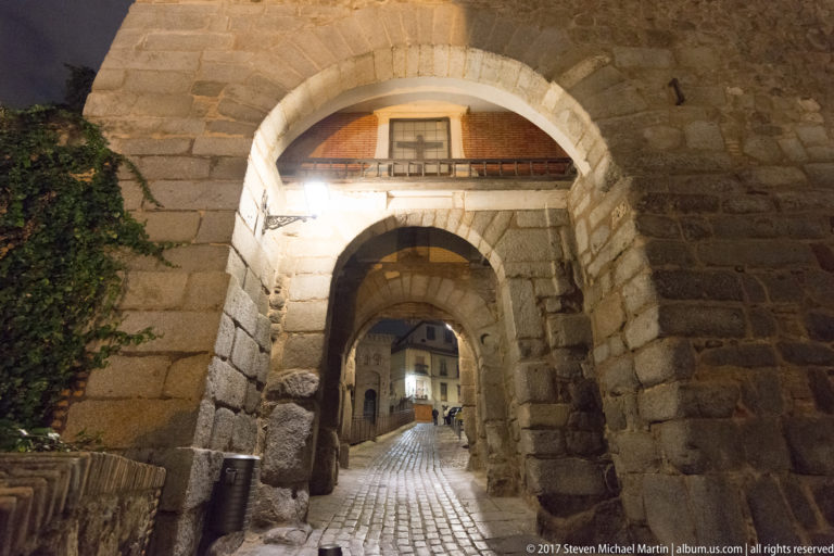 Successive stone arches frame a cobbled passage through a thick fortified city gate in Toledo at night.