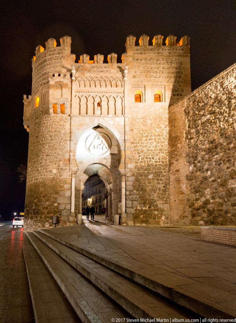 Illuminated Puerta del Sol with horseshoe entrance, flanking towers, crenellations, decorative arches, and a sculpted medallion.