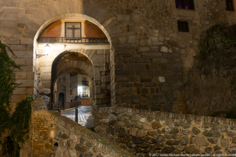 Stone approach to Puerta del Sol with rough fortified walls, layered arches, and a narrow illuminated cobbled passage.